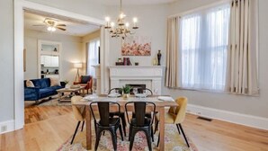 A dining area with a wooden table, black chairs, and a chandelier. There is a fireplace with a mantel and a painting above it. The room has wooden flooring and a large window with curtains. - 4 Units! Mins to Downtown&Dining - Dauphin Darling (Mobile)