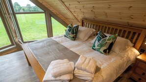 Een slaapkamer met een houten plafond, grote ramen, een bed met witte lakens en uitzicht op een groen veld.