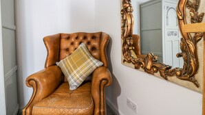 Un fauteuil en cuir marron avec un coussin à carreaux, placé à côté d’un grand miroir décoratif sur un mur blanc.