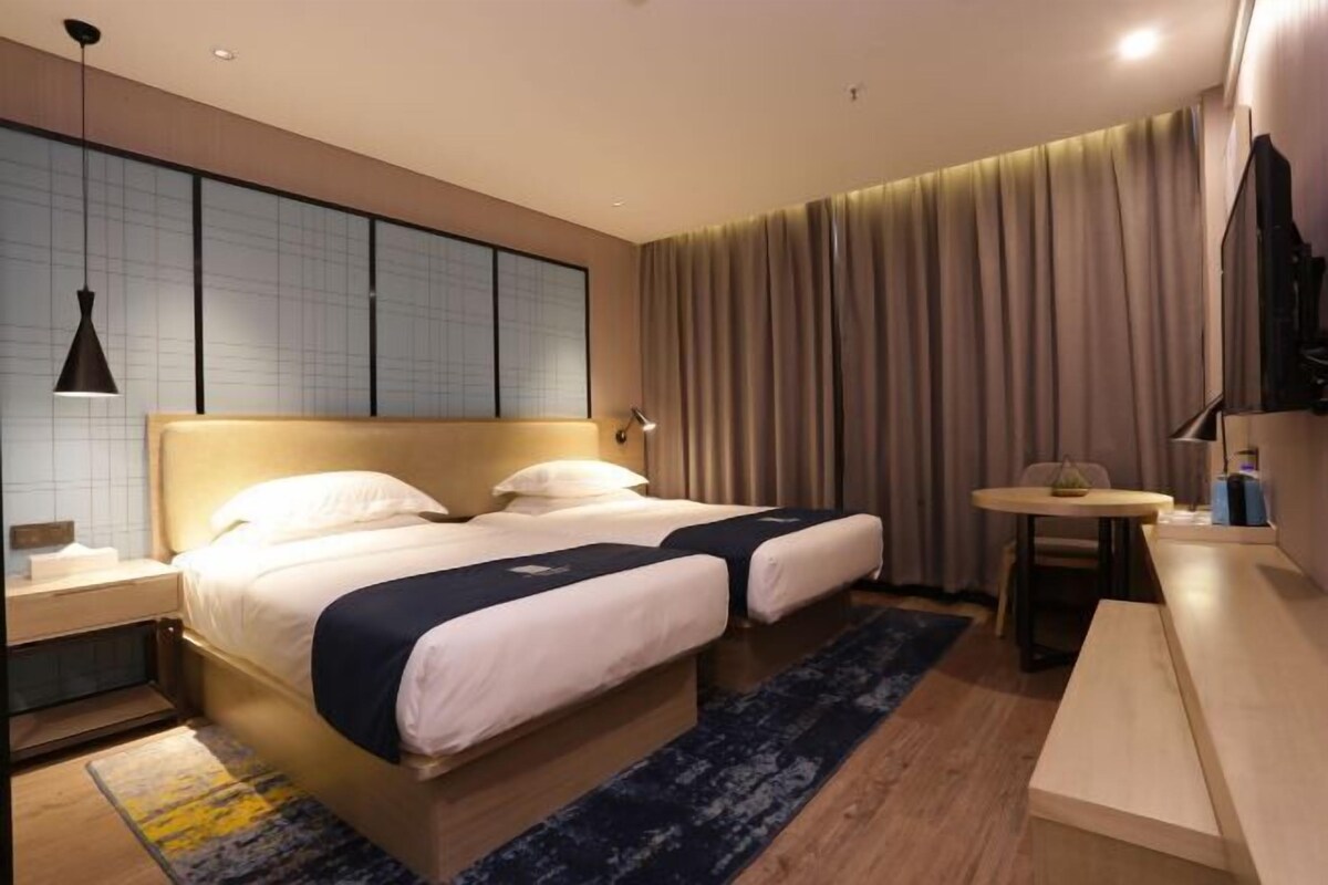 A hotel room with two beds, a small table, a chair, and a large window with curtains.