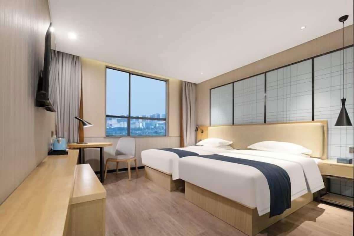 Une chambre d’hôtel avec deux lits, un bureau, une chaise, une télévision et une grande fenêtre donnant sur la ville.