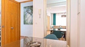Una habitación de hotel con una puerta de madera, una mesa de vidrio con un cuenco, una silla color verde azulado, un sofá y un cuadro en la pared.