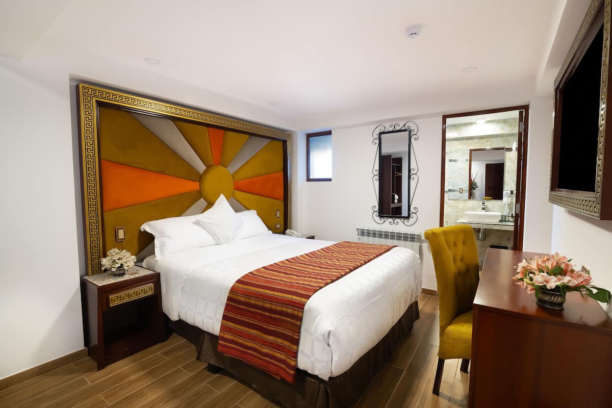 A hotel room with a large bed, a bedside table with a lamp, a chair, a desk with a vase of flowers, and a mirror on the wall.