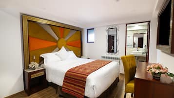 A hotel room with a large bed, a bedside table with a lamp, a chair, a desk with a vase of flowers, and a mirror on the wall.