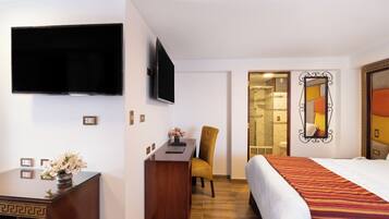 Quarto de hotel com uma cama, uma escrivaninha com cadeira, uma televisão e um banheiro visível por uma porta aberta.