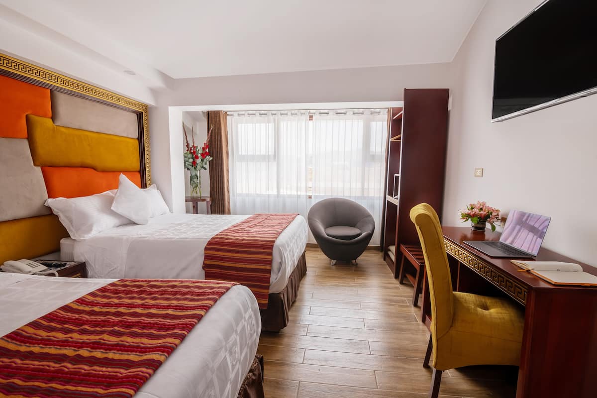 A hotel room with two beds, a desk with a laptop, a chair, and a TV on the wall.