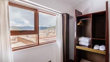 A hotel room with a view of a mountainous landscape through a window. There is a wooden shelf with towels and a pair of slippers.