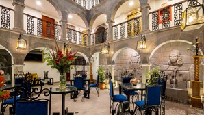 Eine großzügige Lobby mit hohen Decken, gewölbten Säulen aus Stein und aufwendigen Geländern aus schwarzem Metall. Tische und blau gepolsterte Stühle bieten Sitzgelegenheiten.