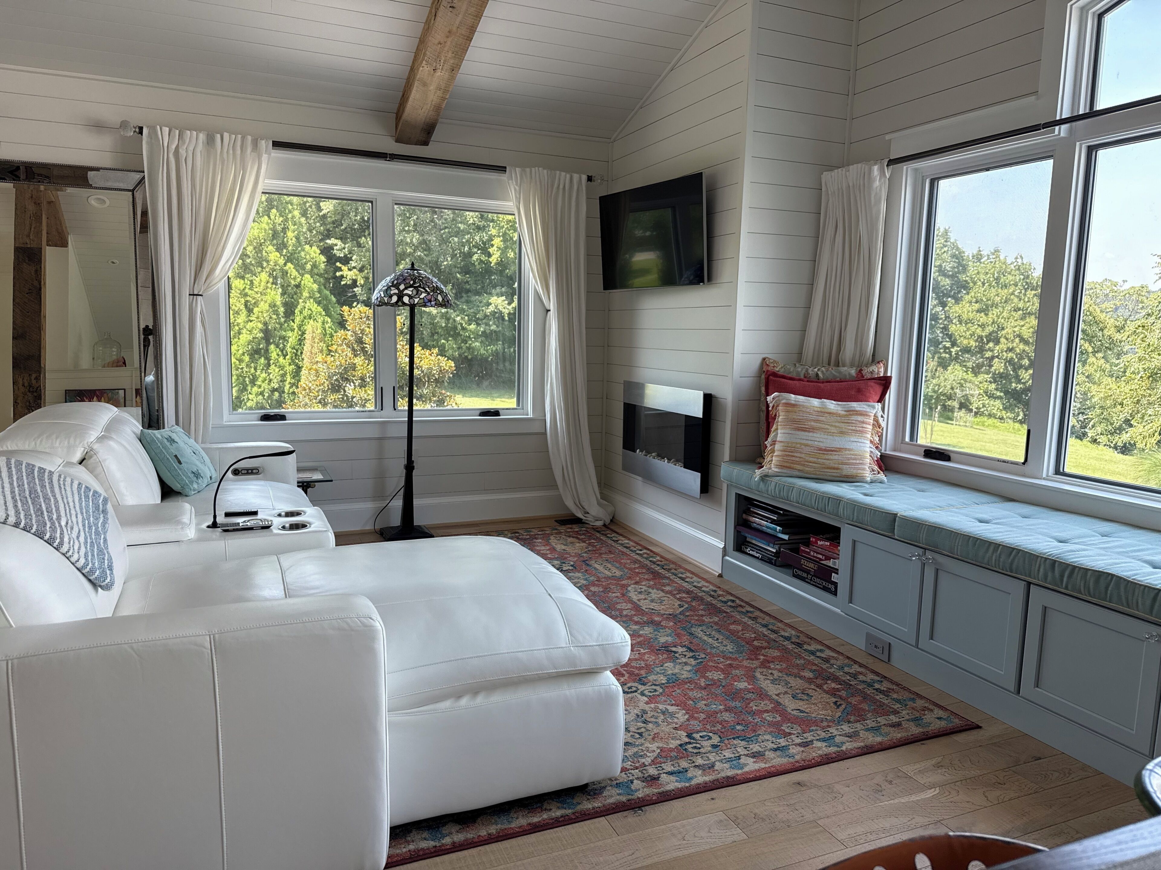 A room with a white leather sofa, a floor lamp, a fireplace, a TV mounted on the wall, and a window with a view of greenery.