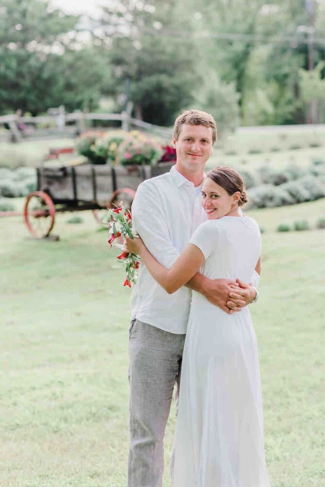A couple embracing in a grassy outdoor area with a cart and flowers in the background.