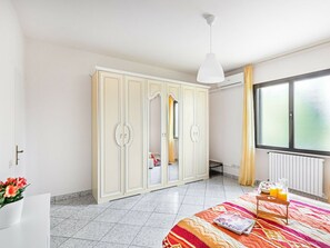 Ein Schlafzimmer mit großem Kleiderschrank, einem Bett mit bunter Bettdecke, einem Nachttisch mit Glas Orangensaft und einem Fenster mit Vorhängen.