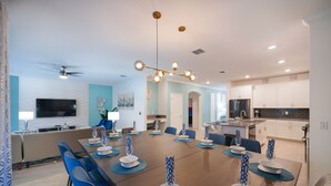 Une salle à manger moderne avec une longue table dressée pour six personnes, des chaises bleues et un lustre.