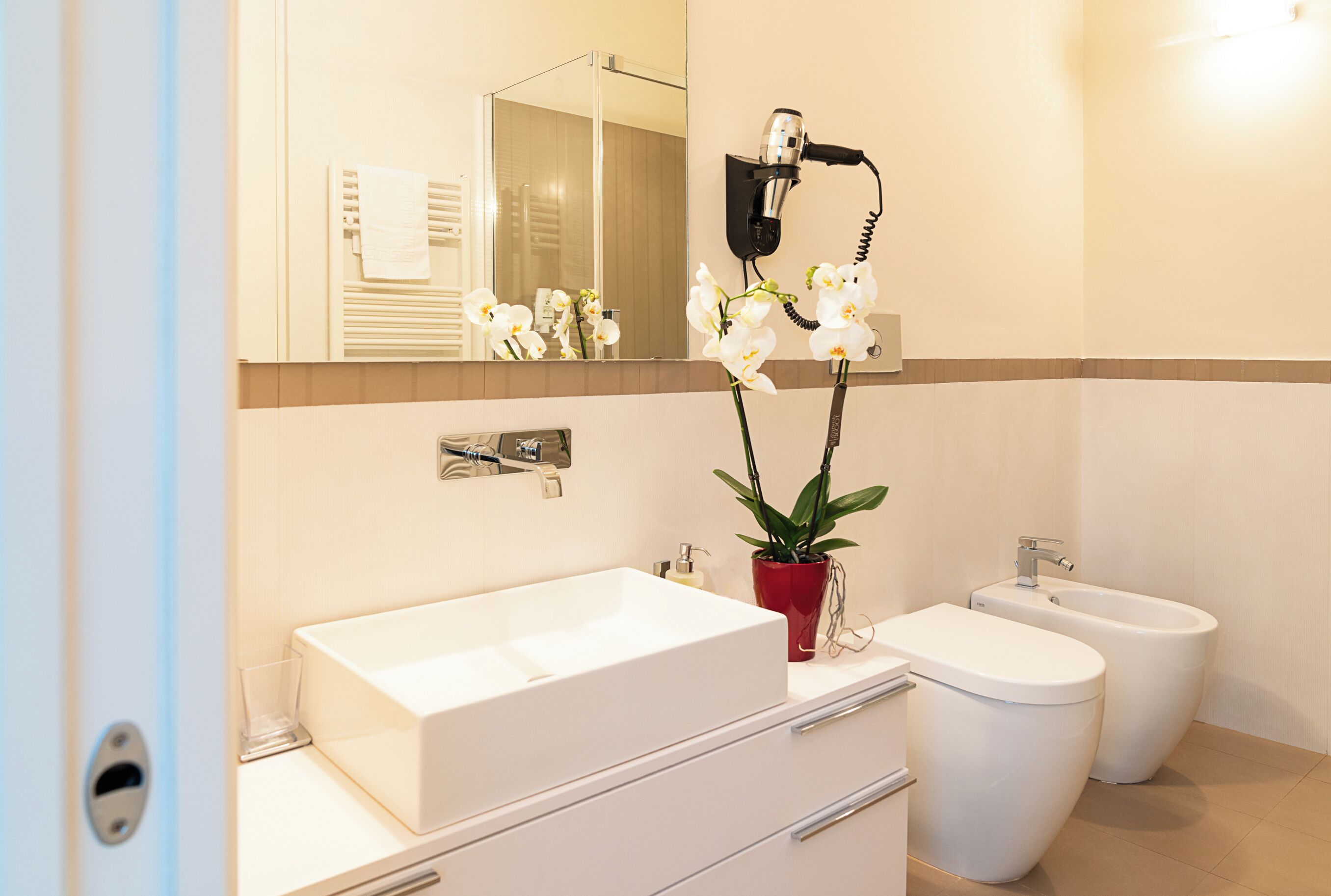 A modern bathroom with a large white sink, a bidet, a hairdryer, and a potted orchid.