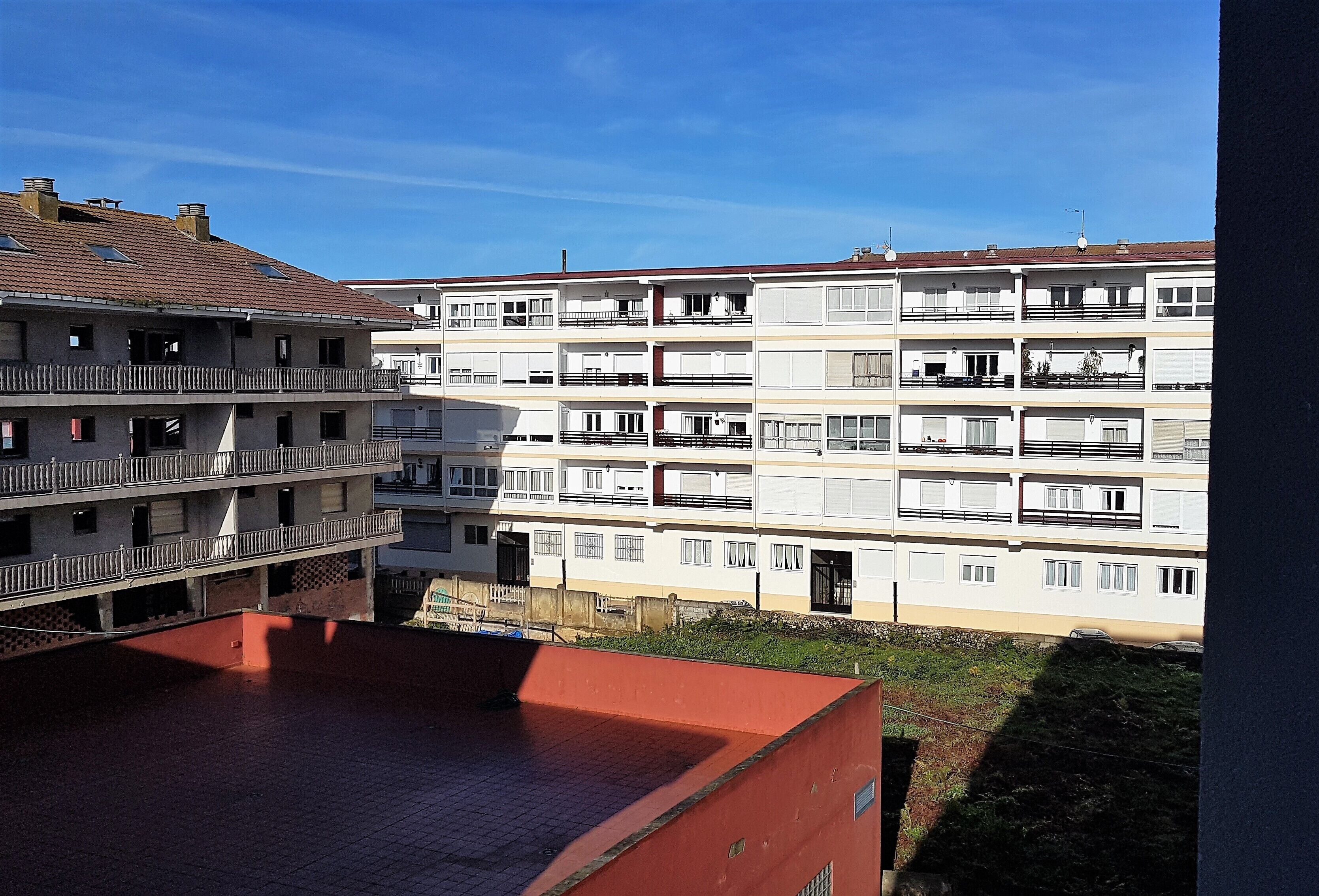 Un edificio residencial de varios pisos con balcones, una construcción con techo rojo en primer plano y un cielo azul despejado.