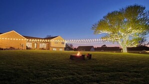Un espace herbeux avec un foyer, un arbre et un bâtiment décoré de guirlandes lumineuses.