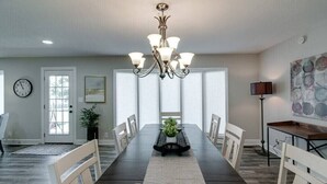 Une salle à manger avec une longue table, des chaises blanches, un lustre, une horloge et une porte avec des fenêtres.
