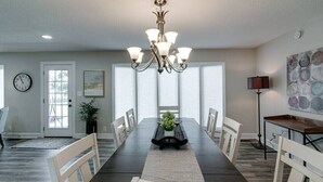 A dining room with a long table, chairs, a chandelier, a clock, a door, a plant, and artwork on the wall.