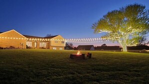 A building with a large open grassy area in front, a fire pit, and string lights.
