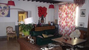 Ein gemütliches Wohnzimmer mit einem grünen Sofa, einem braunen Ledersessel und einem Couchtisch. Eine rote Lampe, ein Topf mit Pflanze und ein blumiger Vorhang sind ebenfalls vorhanden.