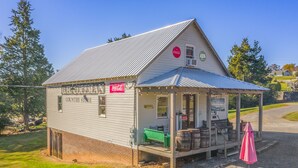 A small, single-story building with a metal roof and a porch. It has a Coca-Cola sign and a sign indicating it is a country store. There are wooden barrels and a green bench outside.