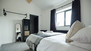En snyggt bäddad säng med vita lakan och en grå filt, ett sängbord med en lampa, en garderob och ett fönster med gardiner.