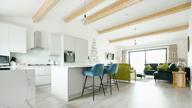 A modern kitchen with white cabinets, a marble backsplash, and a wooden ceiling. There is a small Christmas tree and a dining area with blue chairs.