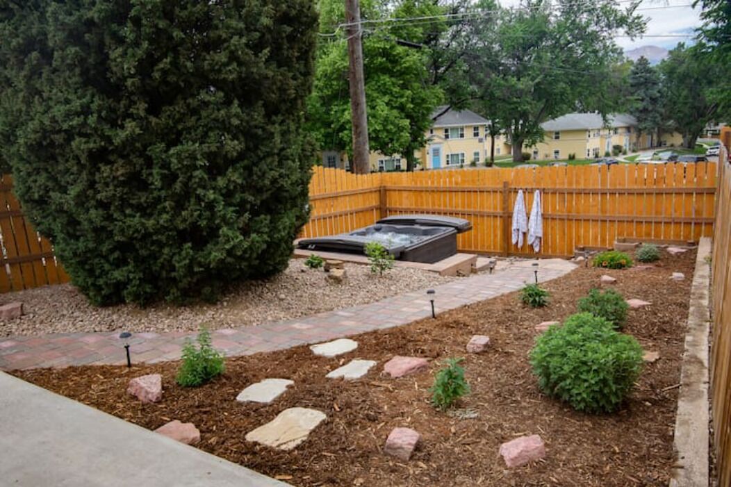 A backyard with a hot tub, a wooden fence, a large tree, and a stone pathway.