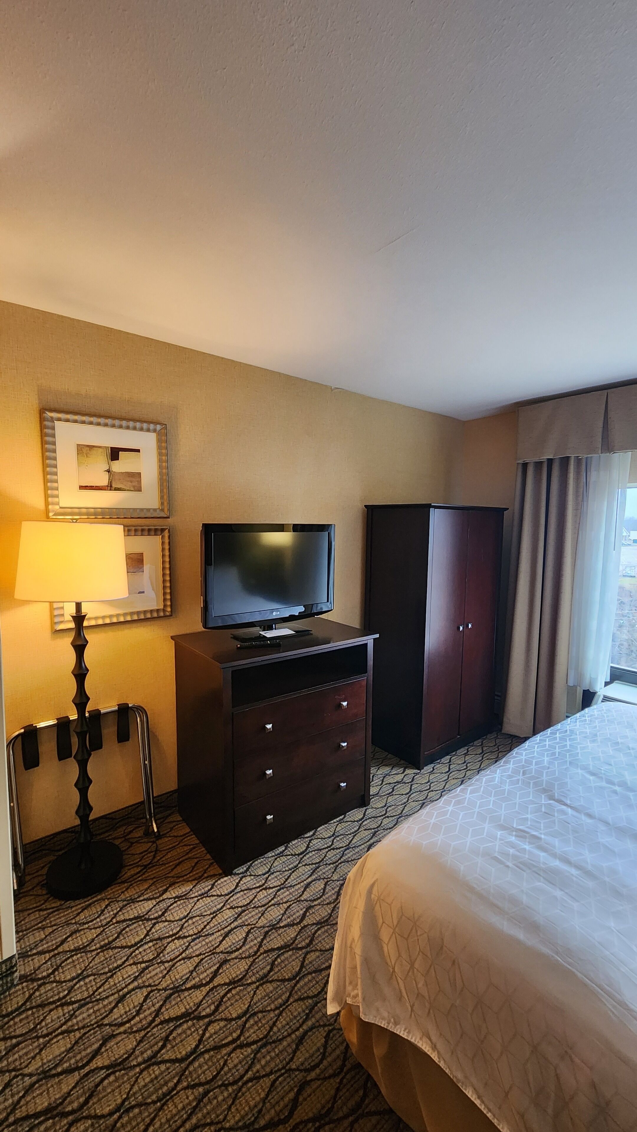A hotel room with a bed, a TV on a dresser, a wardrobe, a lamp, and a framed picture on the wall.