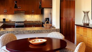 A modern kitchen with dark wooden cabinets, a granite countertop, and a stainless steel refrigerator. There is a round table with a bowl of fruit in the center. - Villas San Ángel (Departamento) (Ajijic)