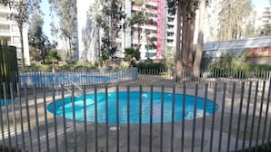 Une piscine entourée d’une clôture métallique, avec une pelouse au premier plan et de grands immeubles en arrière-plan.