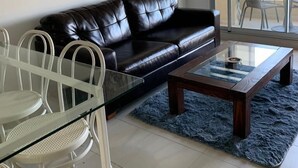 Un salon moderne avec un canapé en cuir noir, une table basse en verre et un coin repas comprenant des chaises blanches et une table en verre.