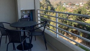 Un balcon avec une table et des chaises, un meuble, et une vue sur un paysage vallonné parsemé de bâtiments.
