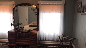 Ein Zimmer mit einem Holzschrank, einem Spiegel und einem kleinen Tisch. Zwei Fenster mit durchsichtigen Vorhängen und ein Teppich auf dem Boden.