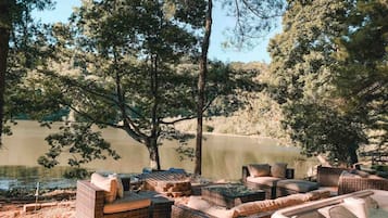 An outdoor seating area with wicker furniture and a hot tub, surrounded by trees and a lake.
