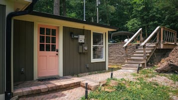 A small, single-story shed with a pink door and a window, surrounded by a brick pathway and greenery.