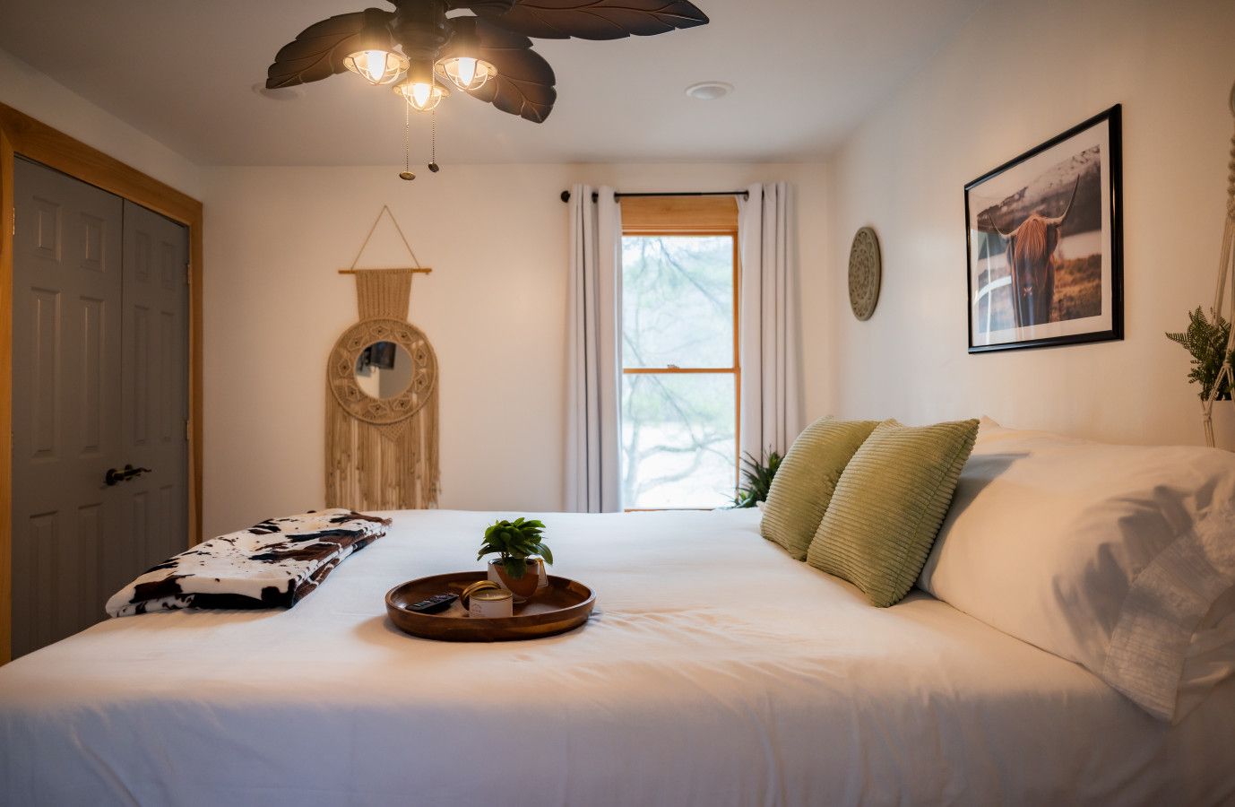 A bedroom with a bed, a bedside table with a tray, a hanging mirror, a window with curtains, and a framed picture on the wall.