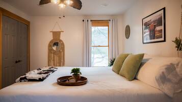 A bedroom with a bed, a bedside table with a tray, a hanging mirror, a window with curtains, and a framed picture on the wall.