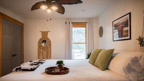 Un dormitorio con cama, mesita de noche con bandeja, espejo colgado, ventana con cortinas e imagen enmarcada en la pared.