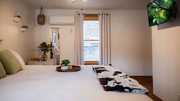 A bedroom with a bed, a TV mounted on the wall, a window with curtains, and a small table with a plant.