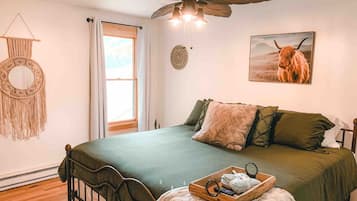 A bedroom with a bed, a ceiling fan, a macrame wall hanging, a window, and a painting of a Highland cow.