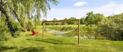 A hammock set up on a wooden frame in a grassy area with a pond and trees in the background.