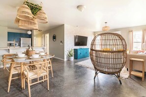 A modern living room with a round dining table, wicker chairs, a large rattan chair, a flat-screen TV, and a kitchen area with blue cabinets and a microwave. - Boho House- A Rural Escape with Fire Pit, EV, WFH (Ithaca)