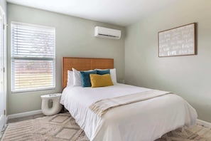 A bedroom with a bed, a window with blinds, an air conditioner, a bedside table, and a wall with a framed text. - Boho House- A Rural Escape with Fire Pit, EV, WFH (Ithaca)