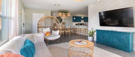 A living room with a sofa, a coffee table, a TV, and a dining area with chairs and a hanging light fixture.