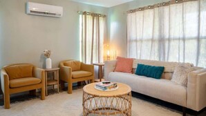A living room with a sofa, two armchairs, a round coffee table, a small side table, and a wall-mounted air conditioner.
