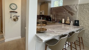A modern kitchen with a marble countertop, wooden chairs, and a stone backsplash.