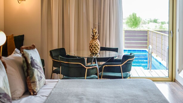 A hotel room with a bed, a glass table with chairs, a pineapple centerpiece, and a view of a pool through a sliding door. - Golden Suites & Spa (Ioannina)