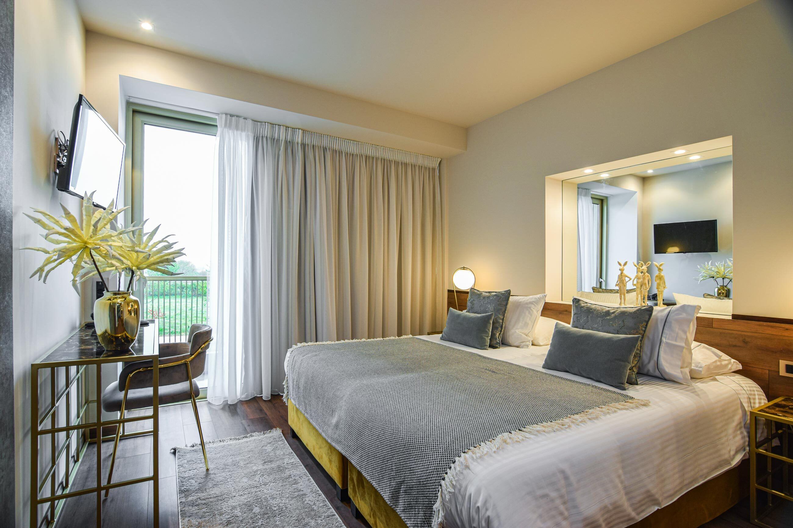 A modern bedroom with a large bed, bedside tables, a desk with a chair, and a view of the outdoors.
