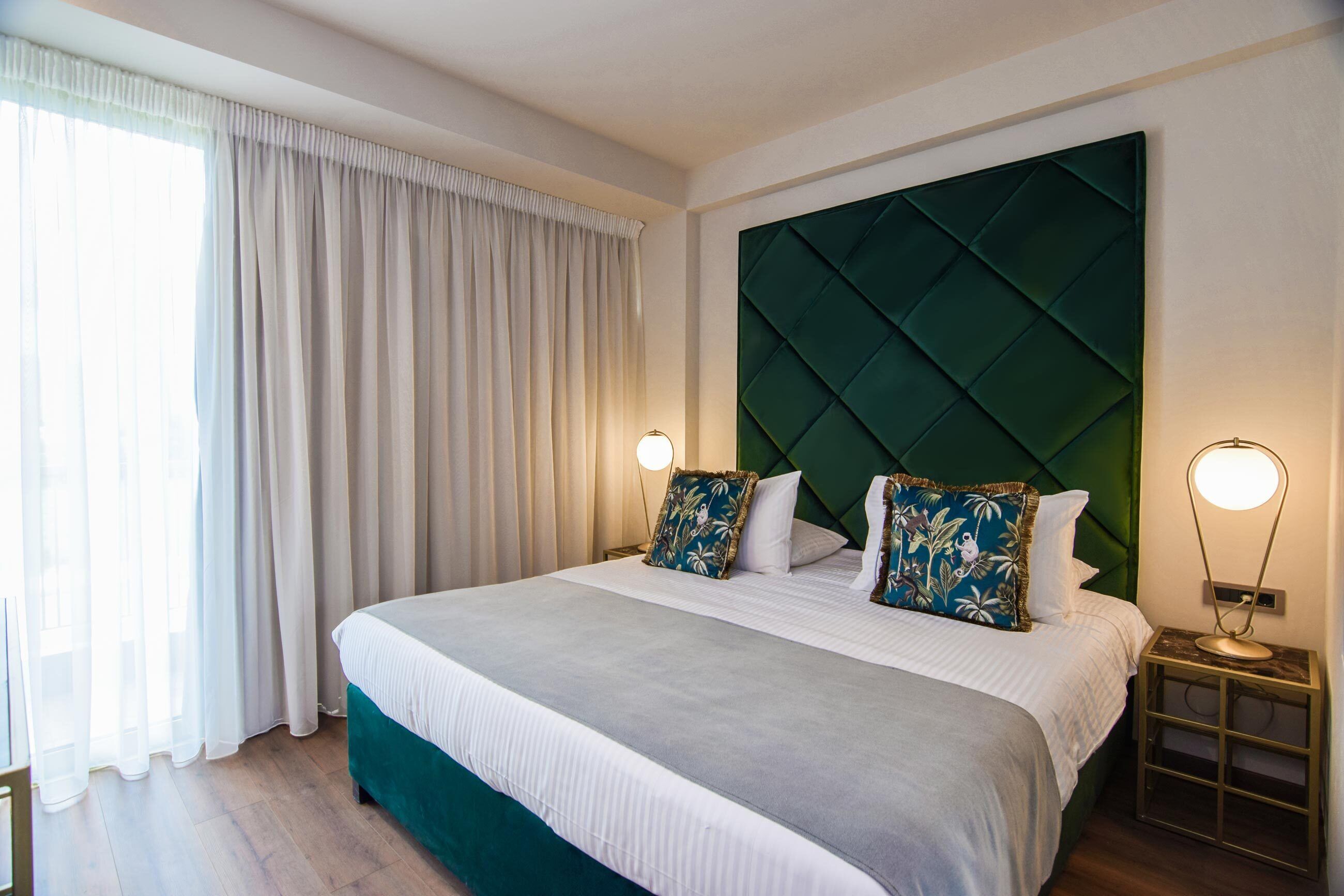 Una cama bien tendida con un cabecero verde, dos lámparas de noche y una ventana con cortinas traslúcidas.