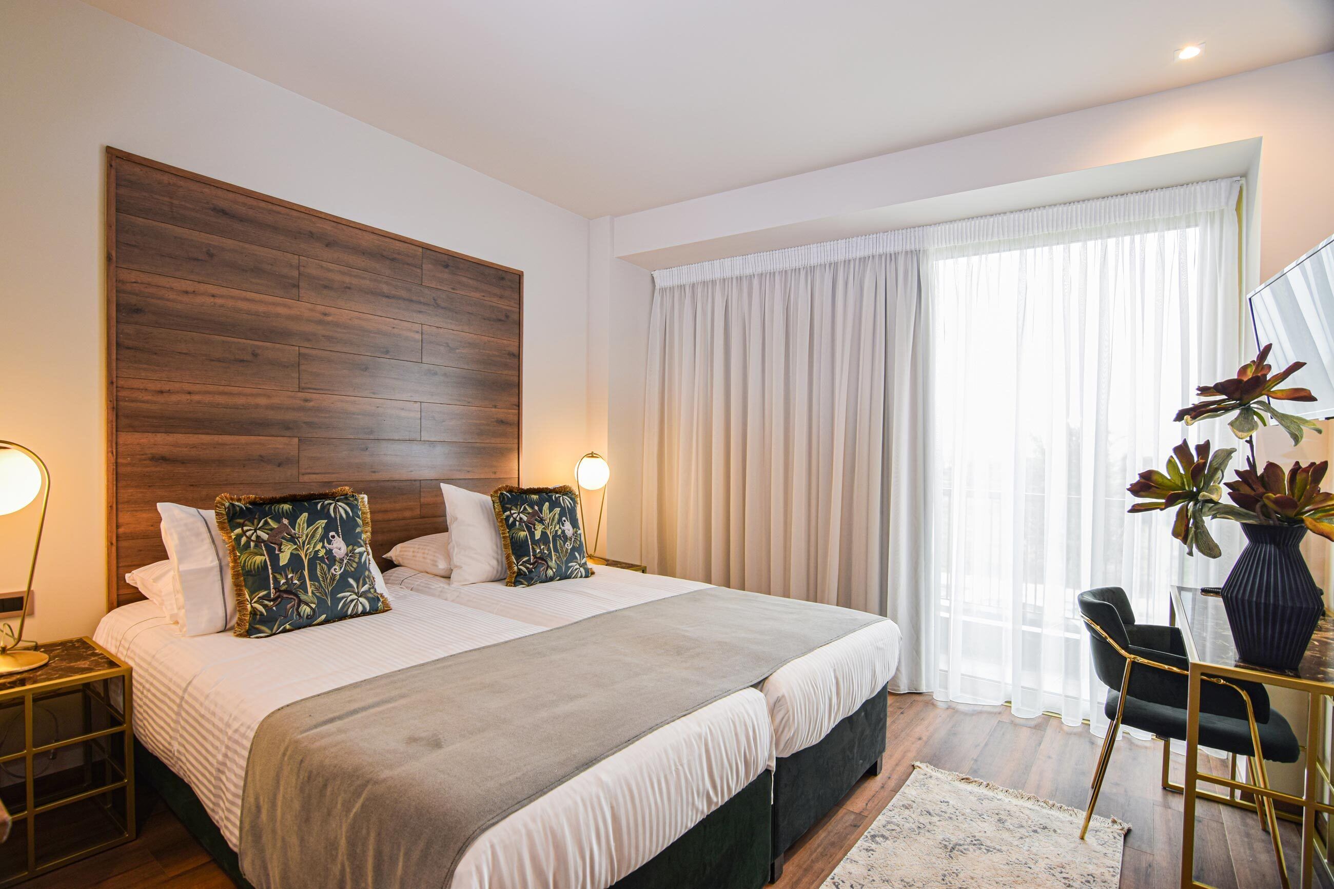 Quarto de hotel com uma cama grande, cabeceira de madeira, escrivaninha com cadeira e janela com cortinas finas.